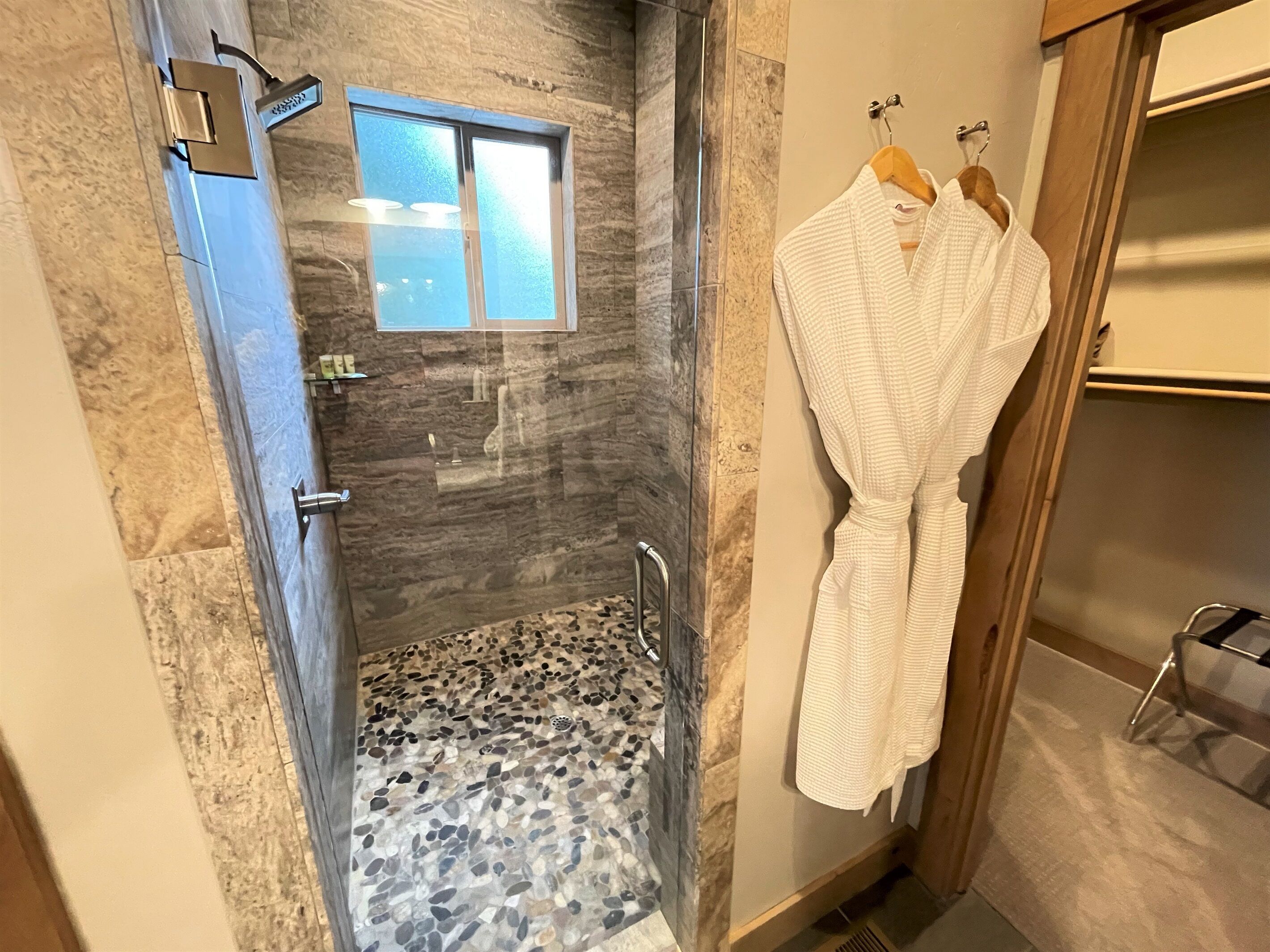 A bathroom with a glass-enclosed shower, a marble floor, and a wooden vanity with a towel rack holding a white bathrobe.