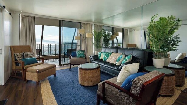 A modern hotel room with a balcony, a sofa set, a dining table, and a large potted plant. - Oceanfront Corner 3BR · Maui Turtle & Whale views (Wailuku)