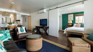 Ein stilvolles Wohnzimmer mit einer Couch, einem Sessel und einem Fernseher. Es gibt einen Essbereich mit Tisch und Stühlen. Der Raum verfügt über einen großen Spiegel und eine Schiebetür zu einem Schlafzimmer mit Bett und Nachttisch.