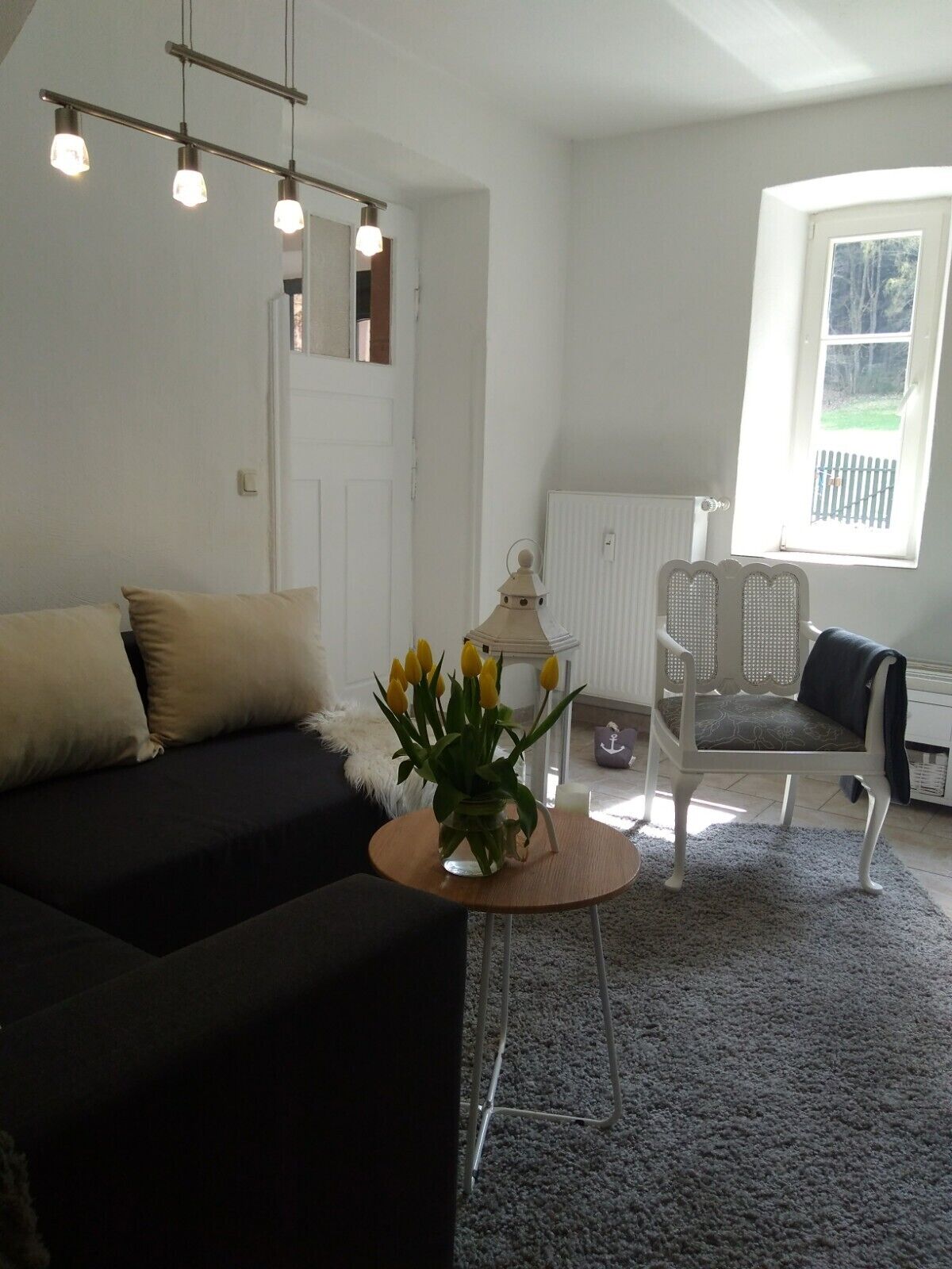Ein modernes Wohnzimmer mit einem dunklen Sofa, einem runden Couchtisch mit einer Vase voller gelber Tulpen und einem weißen Sessel mit einem gemusterten Kissen. Ein großes Fenster bietet Blick auf einen Zaun.