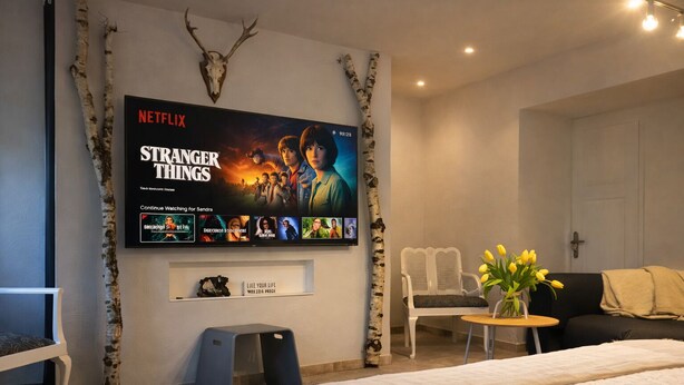 Ein modernes Schlafzimmer mit einem großen Flachbildfernseher an der Wand, der 'Stranger Things' zeigt. Es gibt ein Bett mit weißer Bettdecke, einen kleinen runden Tisch mit einer Vase gelber Blumen und einen weißen Stuhl.