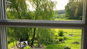 Blick durch ein Fenster auf einen gepflegten Garten mit Schaukel, kleinem Baum und Bank.