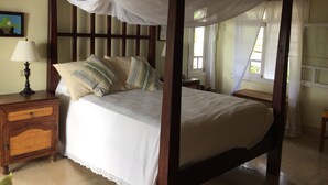 A four-poster bed with a canopy, a wooden nightstand with a lamp, and a wicker cabinet. - Secluded skytop retreat with stunning sea and mountain Views (Falmouth Harbor)