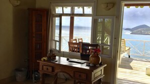 A room with a wooden desk, a chair, a vase with flowers, and a view of the ocean through large windows. - Secluded skytop retreat with stunning sea and mountain Views (Falmouth Harbor)