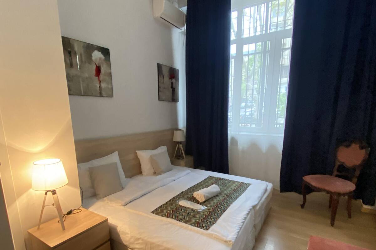 A hotel room with a bed, bedside table, lamp, chair, and artwork on the wall.