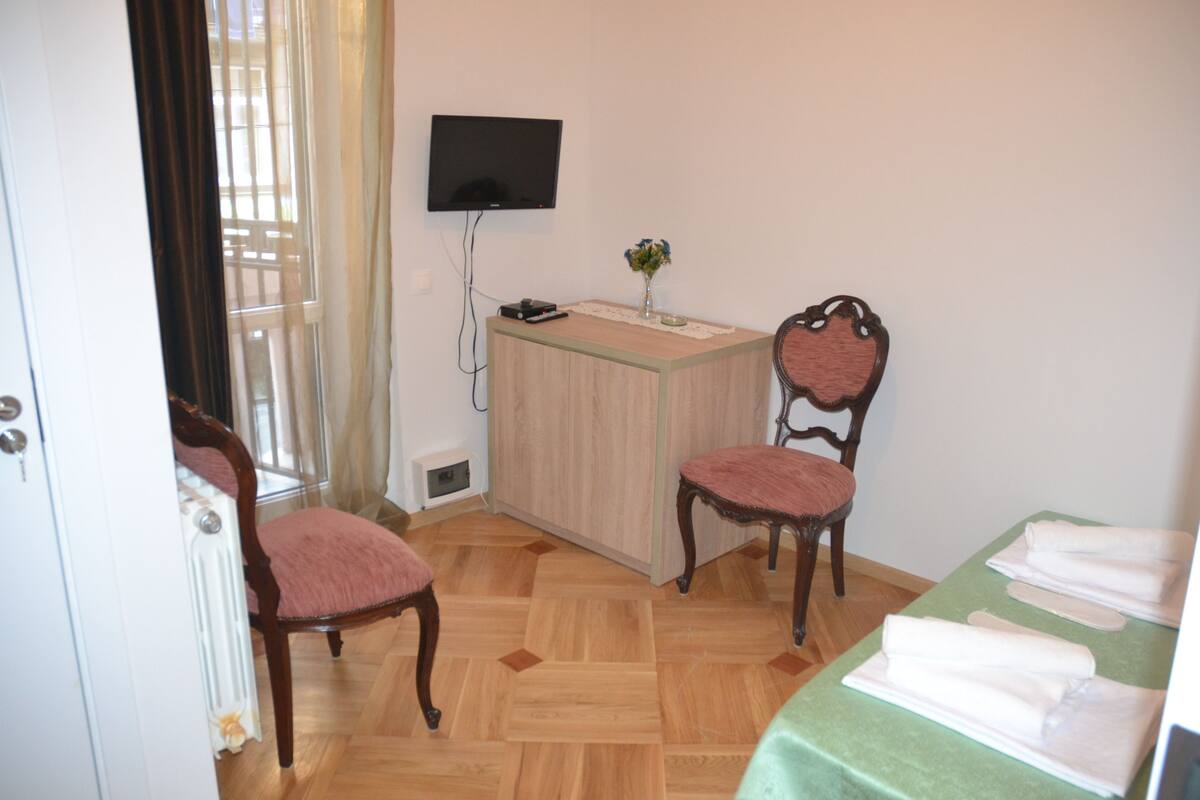 A room with a wooden floor, a desk with a flat-screen TV, two chairs, a small table with a vase, and a bed with towels.