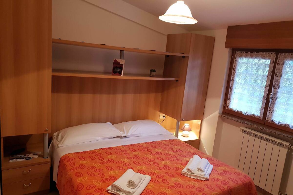 Une chambre à coucher avec un lit, une table de chevet, une étagère avec un livre, une lampe et une fenêtre agrémentée de rideaux décoratifs.