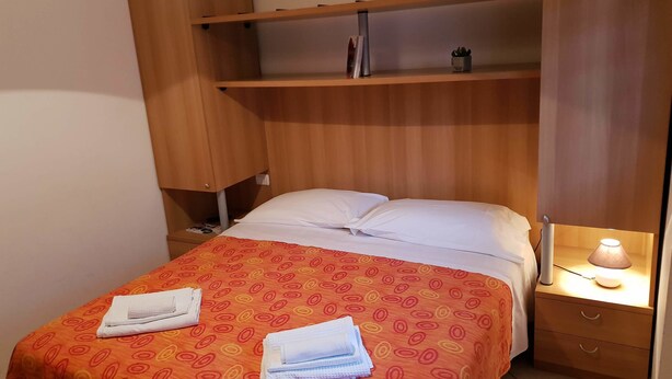 Ein ordentlich bezogenes Bett mit einem gemusterten Überwurf, einer Nachttischlampe und einem kleinen Regal darüber.