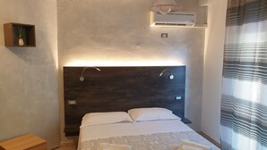 Une chambre moderne avec un grand lit, une table de chevet, une petite étagère avec une plante, un appareil de climatisation et une fenêtre avec des stores.