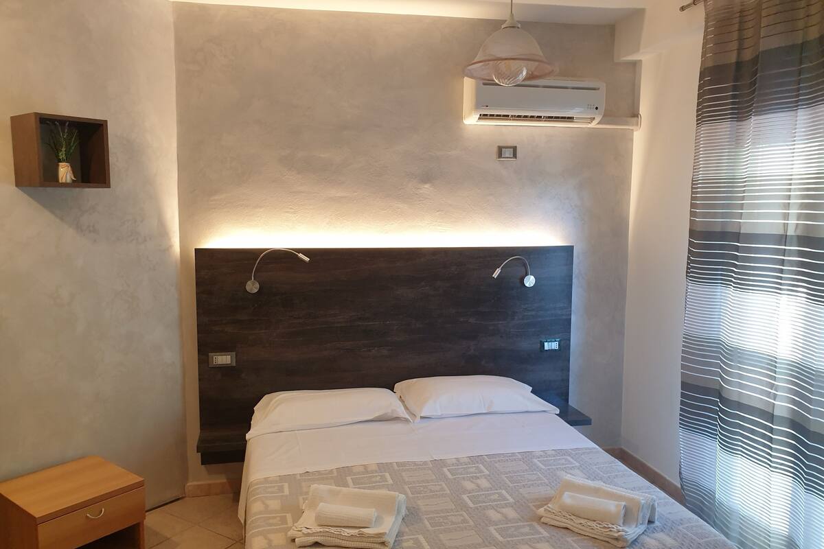 Une chambre moderne avec un grand lit, une table de chevet, une petite étagère avec une plante, un appareil de climatisation et une fenêtre avec des stores.
