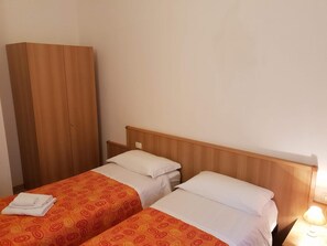 Une chambre d’hôtel avec deux lits simples, chacun doté d’une tête de lit en bois et de linge de lit blanc. On y trouve également une armoire en bois et une table de chevet avec une lampe.