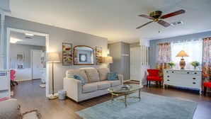 Ein Wohnzimmer mit einem beigefarbenen Sofa, einem Glastisch, einem roten Sessel, einer weißen Kommode und einem Deckenventilator.