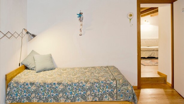 A bedroom with a wooden bed, a patterned bedspread, a wall-mounted lamp, and a small decorative item.