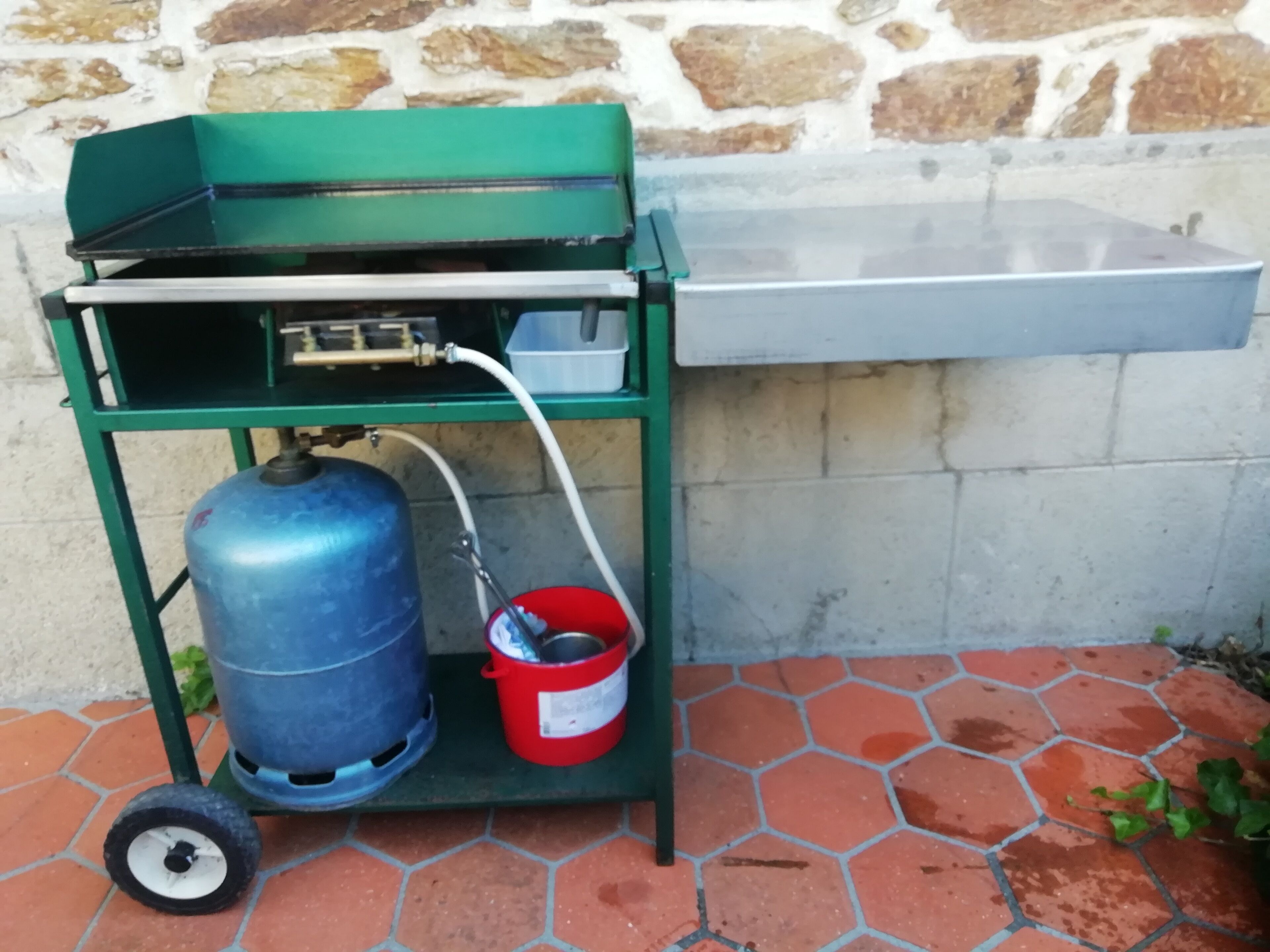 Une bouteille de gaz portable, munie d’un seau rouge fixé à son sommet, est placée sur un chariot métallique vert, à côté d’un mur de pierre.