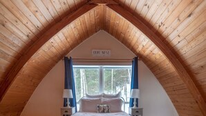 Un dormitorio acogedor en el desván con techo de madera inclinado, una cama grande y una ventana con cortinas.