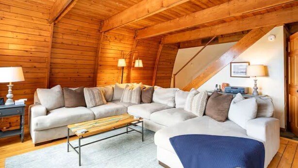 Acogedor interior de cabaña de madera con un sofá seccional, una mesa de centro y una escalera que sube a un nivel superior.