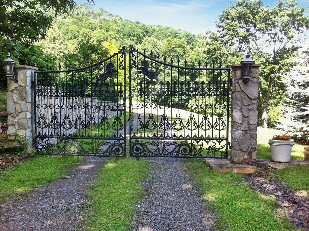 A wrought iron gate with decorative elements, flanked by stone pillars and a pathway leading through a landscaped area.