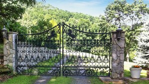 Un portail en fer forgé agrémenté d’éléments décoratifs, flanqué de piliers en pierre et d’un chemin menant à travers un espace paysager.