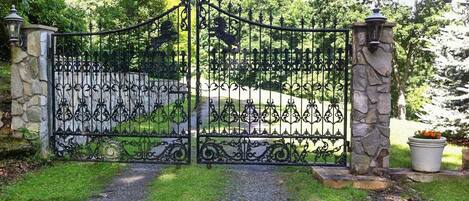 A wrought iron gate with decorative elements, flanked by stone pillars and a pathway leading through a landscaped area.