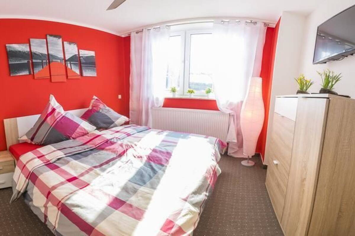 A bedroom with a red accent wall, a bed with a checkered bedspread, a nightstand, a dresser, a flat-screen TV, and a window with curtains.