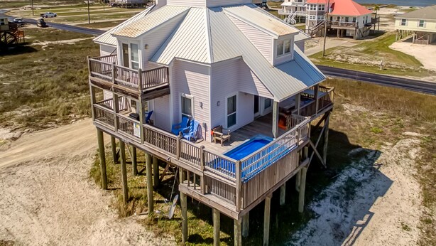 Una casa de madera de varios niveles con piscina en el segundo nivel, construida sobre pilotes sobre una playa arenosa.
