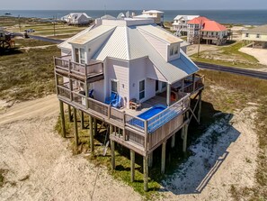 Une maison en bois à plusieurs niveaux, dotée d’une piscine au deuxième étage, construite sur pilotis au-dessus d’une plage de sable.
