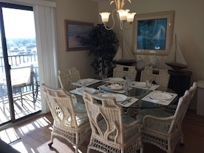 A dining area with wicker chairs and a glass table, framed artwork on the walls, and a view through a sliding door.