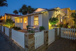 Una casa de dos pisos con cerca blanca, muros de piedra y un porche delantero bien iluminado con mobiliario exterior.