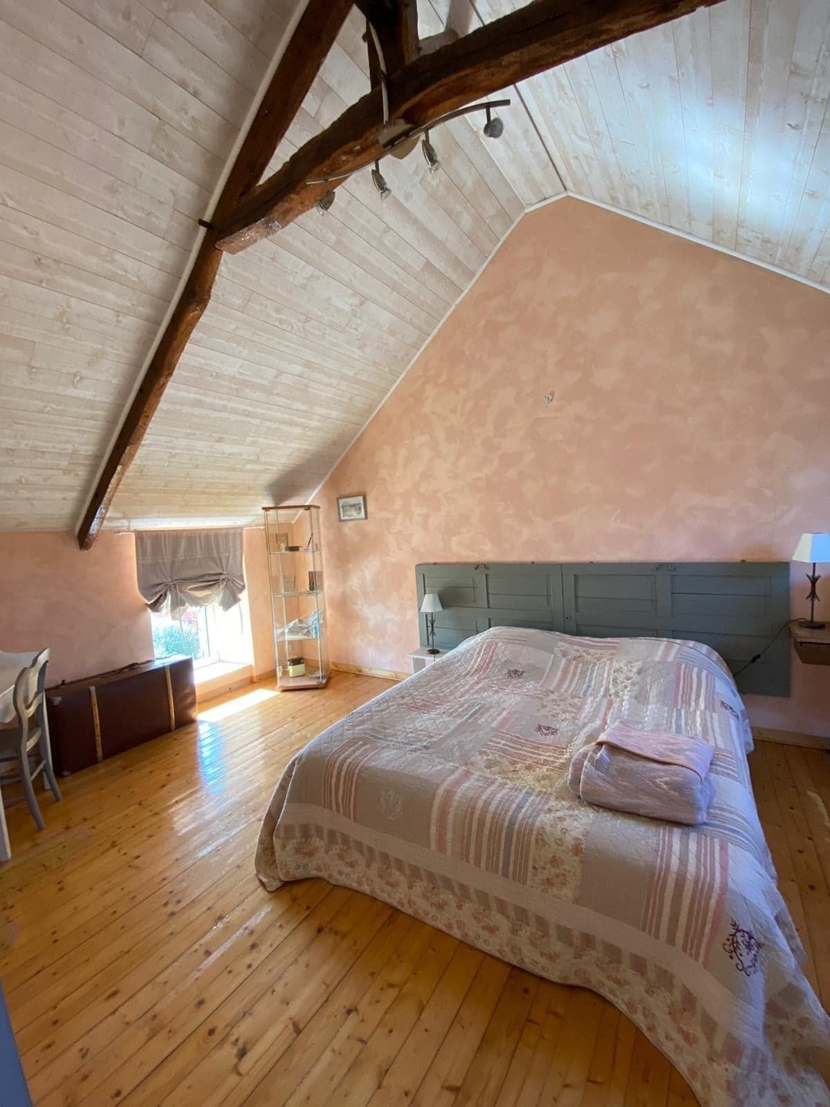 Een slaapkamer met een schuine kap, een bed met een beddengoed met een patroon, een nachtkastje, een stoel en een raam met gordijnen.