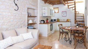 Ein gemütliches Wohnzimmer mit einer Couch, einem Couchtisch und einem Essbereich mit Stühlen und einem Tisch. Im Hintergrund ist eine Küche mit weißen Schränken und einer Wendeltreppe zu sehen.