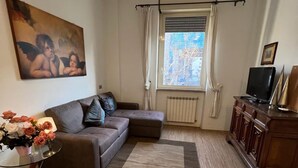 Olohuoneessa on ruskea sohva, lasinen sohvapöytä, jossa on kukkia, suuri taulu seinällä, ikkuna verhoilla ja televisio puukehikkoisessa kaapissa.