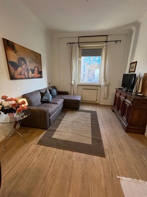 A living room with a brown sofa, a glass coffee table with flowers, a large painting on the wall, a window with curtains, and a television on a wooden cabinet.