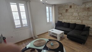 A living room with a stone wall, a dark sofa, a white coffee table, and a glass table with woven placemats. - Renovated, two-room flat with air-conditioned, Vernet district, in city centre (Avignon)
