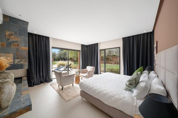 A modern bedroom with a large bed, a stone accent wall, a seating area with chairs, and a view of the outdoors.
