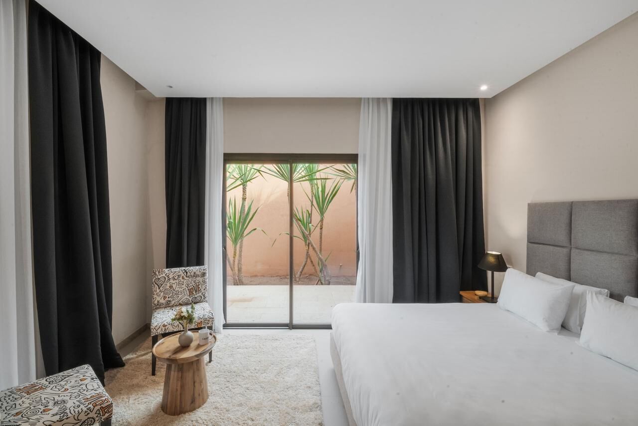 Une chambre d’hôtel avec un lit, une chaise, une petite table et une vue sur les palmiers, visible à travers une porte vitrée.