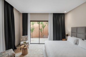 Une chambre d’hôtel avec un lit, une chaise, une petite table et une vue sur les palmiers, visible à travers une porte vitrée.
