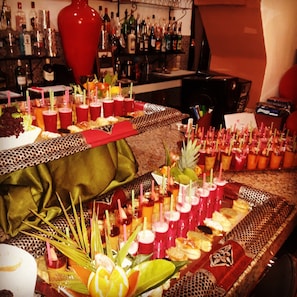 A buffet table with various food items and drinks, a large red vase, and a bar area with bottles in the background. - Villa 865 + (Marrakech)