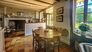 Une cuisine traditionnelle avec une cheminée, une table à manger en bois et des chaises. Une fenêtre offre une vue sur la verdure à l’extérieur.