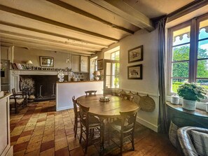 Een traditionele keuken met een open haard, een houten eettafel en stoelen. Er is een raam met uitzicht op groen.
