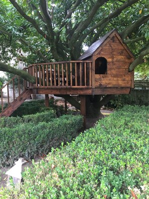 Een houten vogelhuisje met een klein deuurtje, een houten trap die erheen leidt, en een omliggende tuin met struiken.
