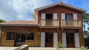 Ein zweistöckiges Holzhaus mit einem Balkon im oberen Geschoss, großen Fenstern und einem Ziegeldach.