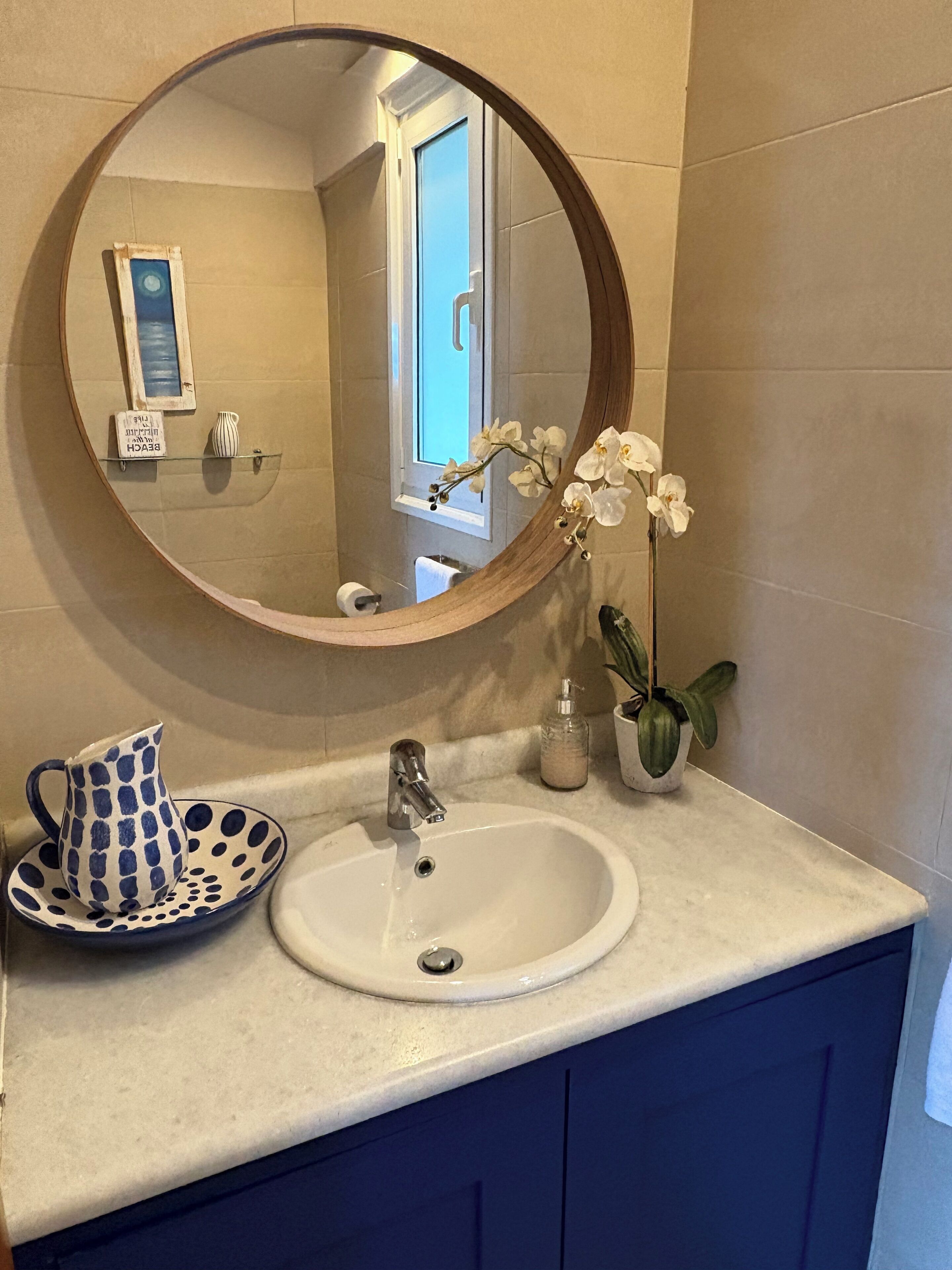A bathroom with a round mirror, a sink, a pitcher, a vase with flowers, and a potted plant.