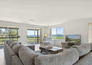 Un salon spacieux avec un grand canapé gris, une table basse, un téléviseur et une vue sur l’extérieur grâce à de grandes fenêtres.