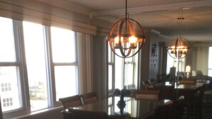 A dining area with a large table and chairs, two pendant lights, and large windows with curtains. - Beautiful Daytona Ocean-Front Condo-Everything You Can Imagine (Daytona Beach Shores)