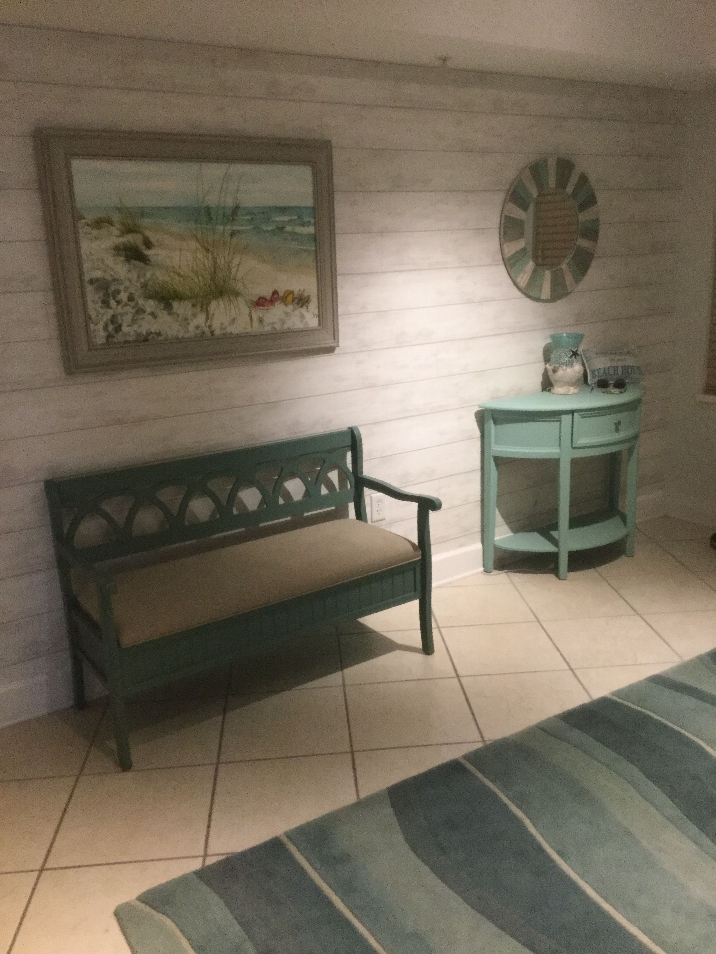 Ein Zimmer mit einer grünen Bank, einem kleinen, türkisfarbenen Beistelltisch, einem Bilderrahmen mit einer Strandansicht und einem dekorativen Wandspiegel.