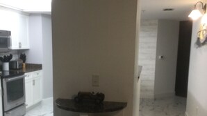 A kitchen with white cabinets, a microwave, and a built-in oven. There is a white shelf unit with multiple shelves, and a dark countertop. - Beautiful Daytona Ocean-Front Condo-Everything You Can Imagine (Daytona Beach Shores)