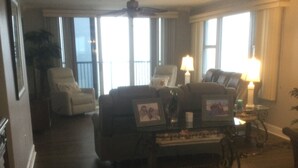 A spacious living room with a dark wood floor, a white armchair, a sofa, and a coffee table with a framed picture. There are two lamps on side tables and a ceiling fan. - Beautiful Daytona Ocean-Front Condo-Everything You Can Imagine (Daytona Beach Shores)