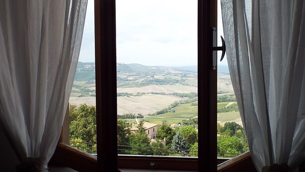 A window with white curtains, revealing a rural landscape with fields and a few buildings.