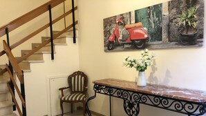 A staircase with wooden railings, a chair with striped upholstery, a marble console table with a vase of white flowers, and a wall-mounted painting of a red scooter.