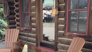 Una cabaña de troncos con dos sillas Adirondack de madera en una terraza de madera, una escoba apoyada contra la pared y un ventanal con marco rojo.